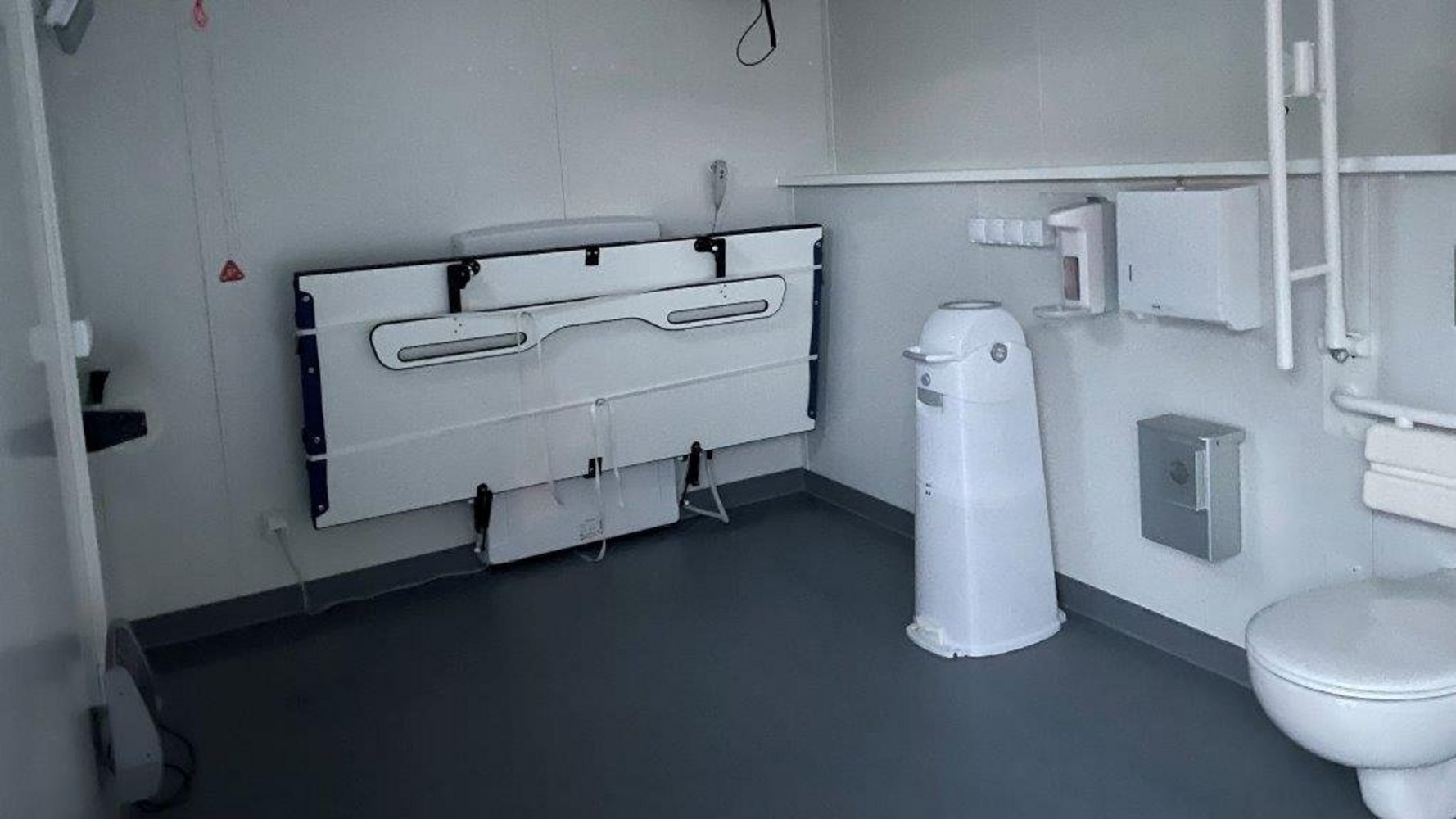 Innenansicht der Toilette für alle mit einer eingeklappten Liege, einem WC und einem Windeleimer.
