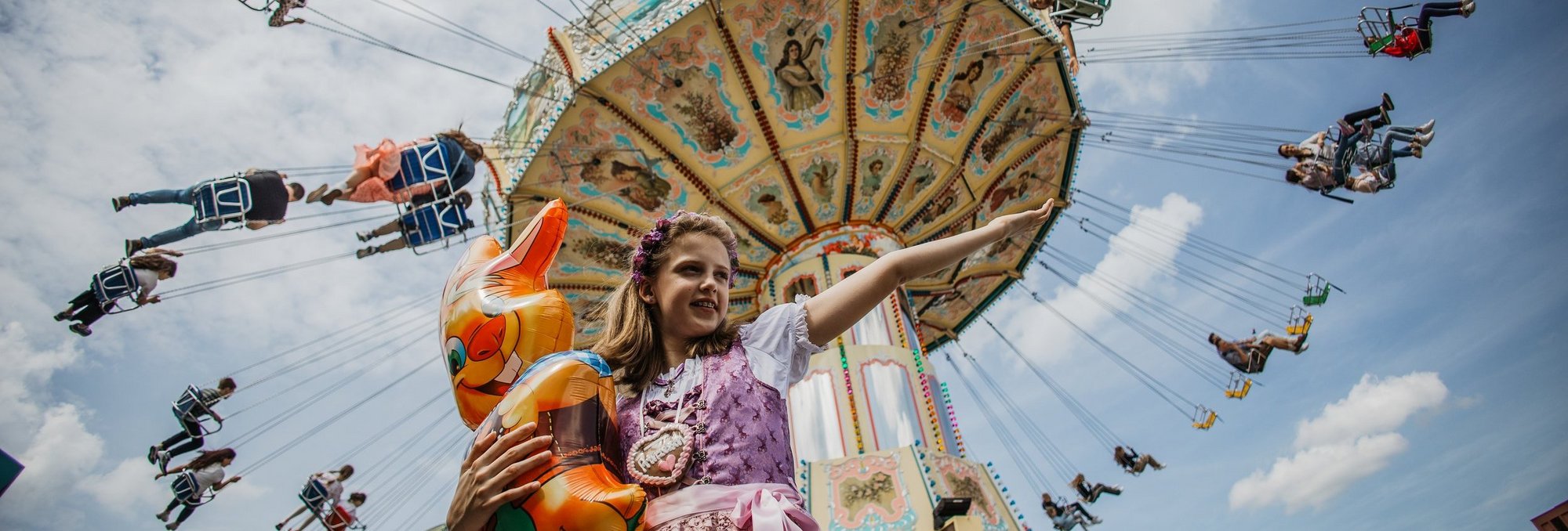 Mädchen mit einem Hasen-Luftballon im Arm steht vor einem Kettenkarussell und streckt den Arm zur Seite aus, so als würde sie das Kettenkarussell präsentieren