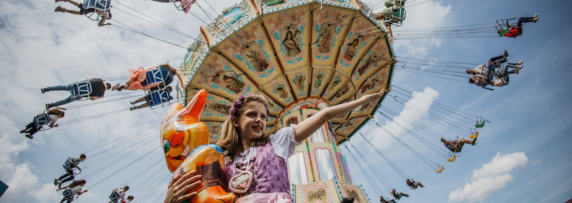 A girl holding a rabbit balloon stands in front of a chain carousel and stretches her arm out to the side, as if she were presenting the chain carousel.
