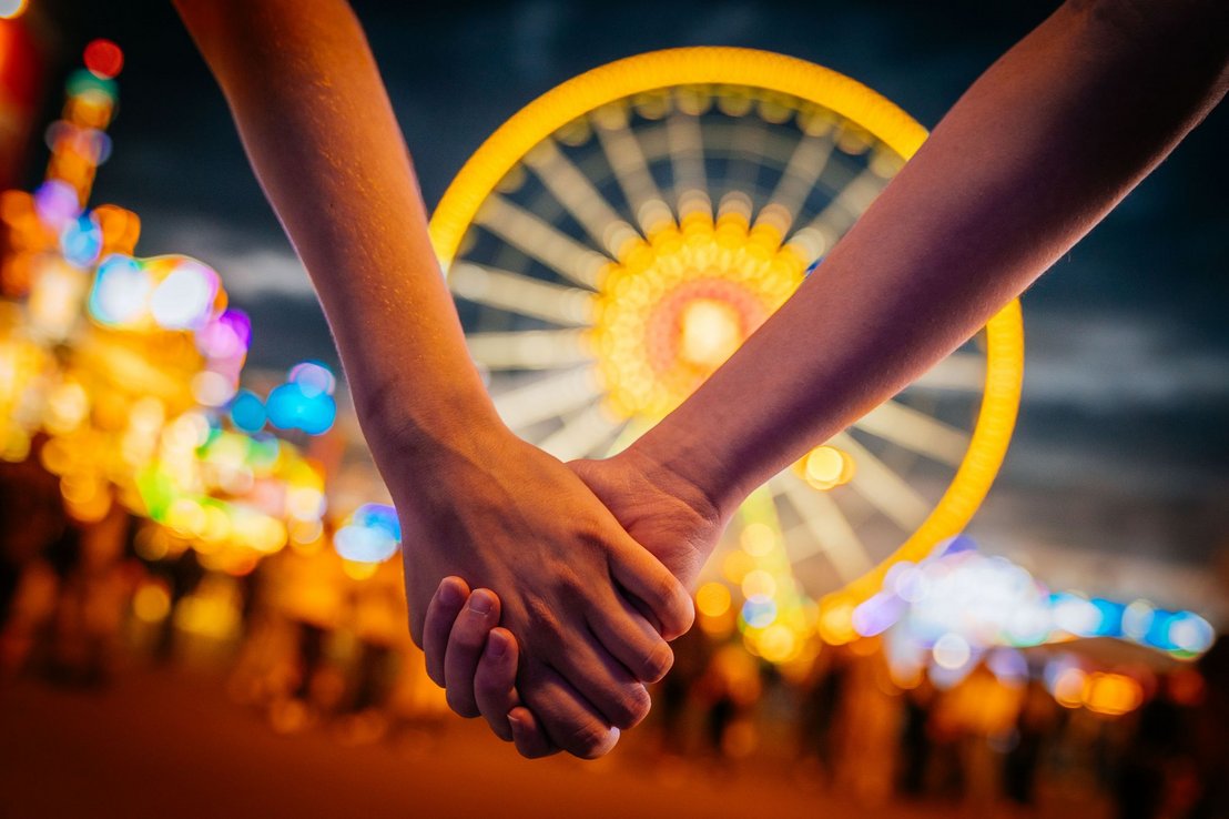 Zwei Menschen halten sich in der Dämmerung an der Hand. Im Hintergrund steht unscharf das beleuchtete Riesenrad.