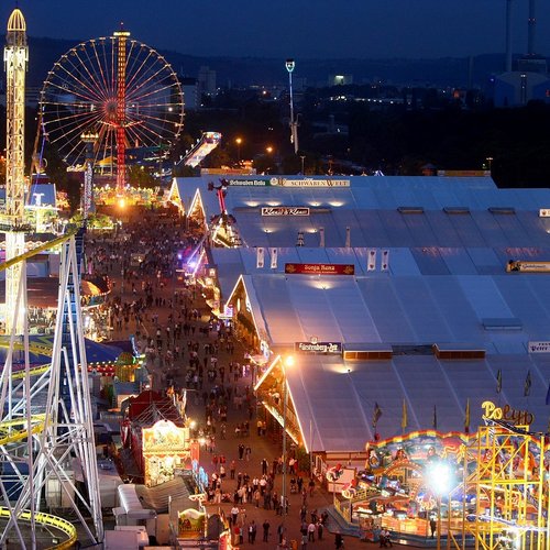 Blick über das Volksfest in der Dämmerung aus der Vogelperspektive. Rechts sind die Festzelte, links lauter Fahrgeschäfte. In der Ferne sieht man das Riesenrad.
