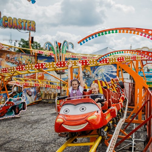 Zwei Kinder fahren in einer Achterbahn, der Wagen sieht aus wie ein roter Rennwagen. Die beiden Kinder jubeln und strahlen.