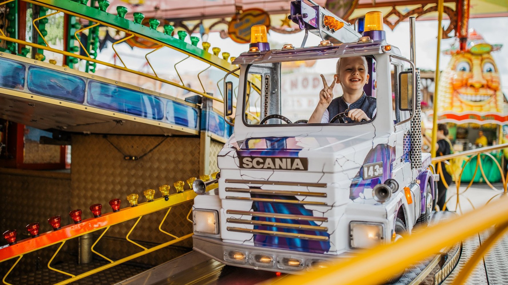 A boy sits in a child's seat in a lorry and makes a peace sign with his fingers towards the camera.