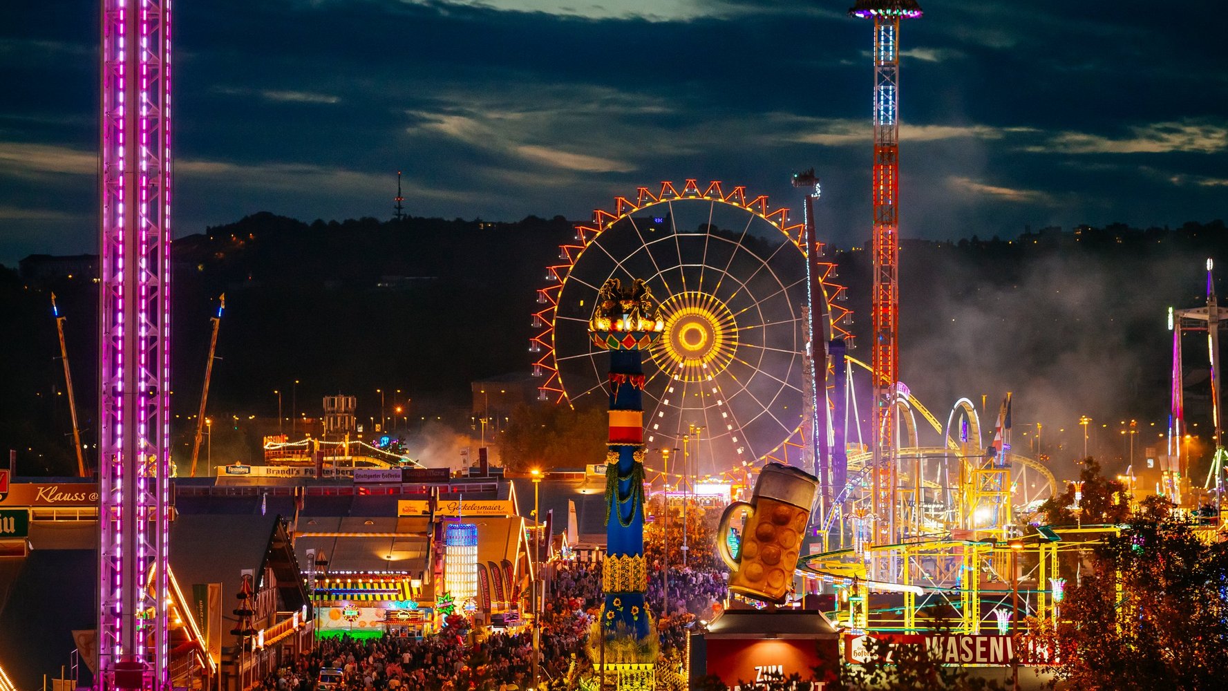 Vogelperspektive auf das Volksfest bei Dämmerung mit vielen beleuchteten Fahrgeschäften und unzähligen Menschen in den Gassen.