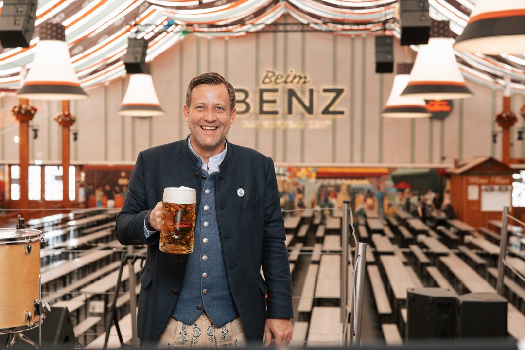 Festwirt Marcel Benz steht auf der Mittelbühne, lächelt in die Kamera mit einem Maßkrug von Stuttgarter Hofbräu in der Hand. 