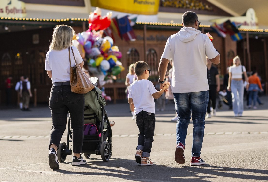 Eine Familie läuft über das Volksfest. Die Frau schiebt einen Kinderwagen, der Mann hält die hand eines Jungen.