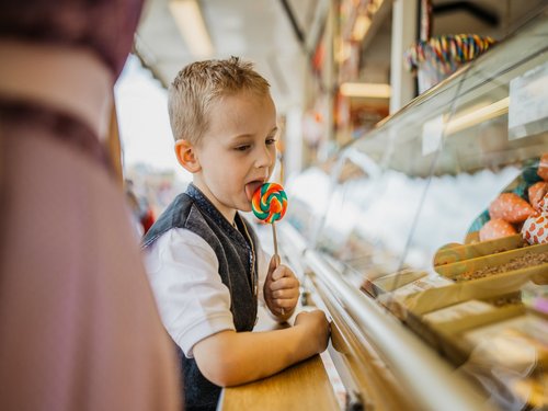 Ein Junge schleckt an einem großen Lolli, während er in eine Süßwaren-Auslage schaut.
