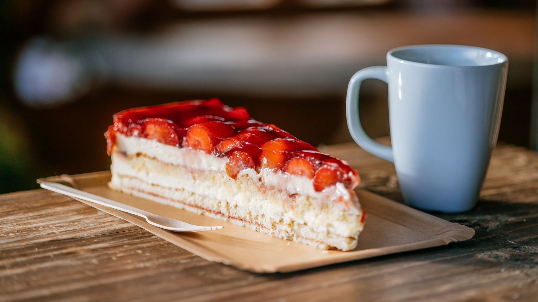 Ein großes Stück Erdbeerkuchen steht auf einem Holztisch. Daneben ist eine Kaffeetasse.