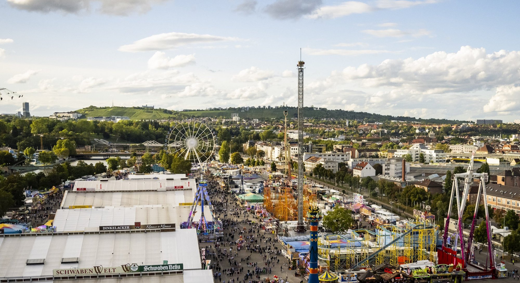 Vogelperspektive auf das Volksfest. Links stehen die Bierzelte, viele Menschen laufen über die Wege. Auf der rechten Seite stehen die Fahrgeschäfte.