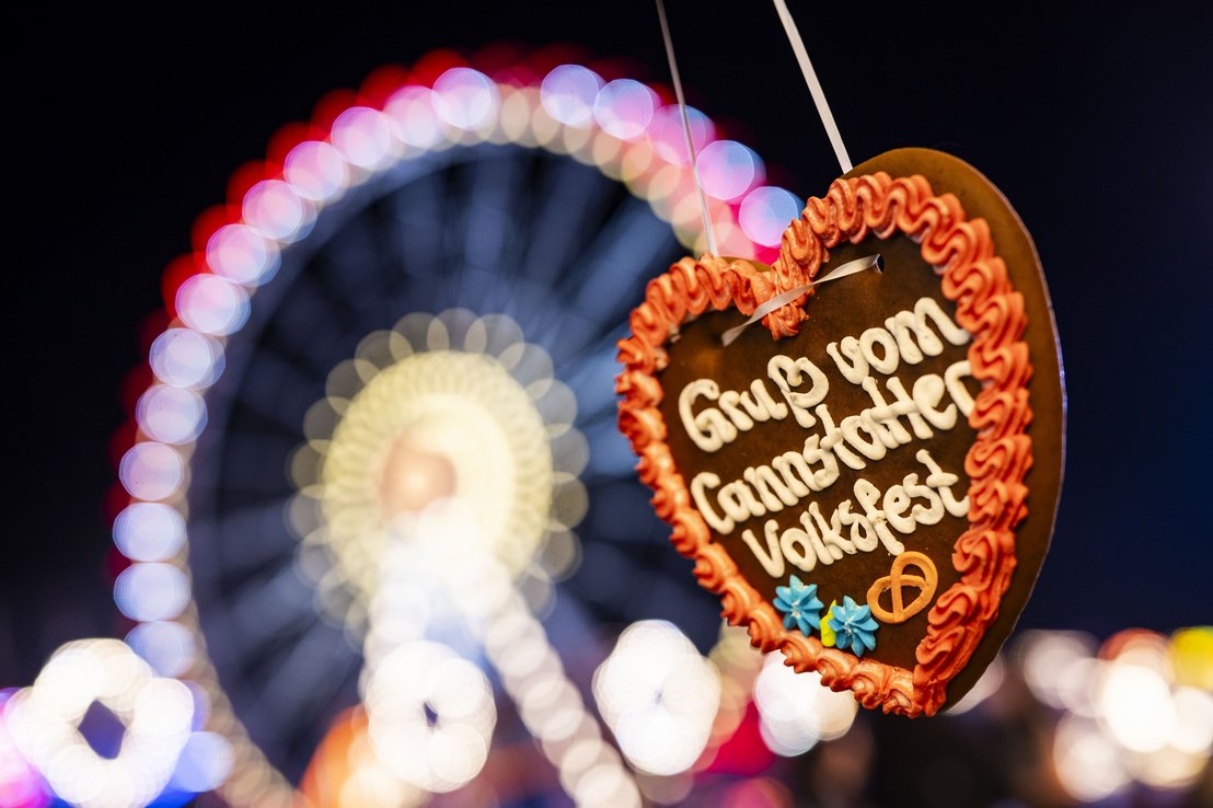 Ein Lebkuchenherz mit der Aufschrift "Gruß vom Cannstatter Volksfest". Im Hintergrund das im Dunklen beleuchtete Riesenrad.