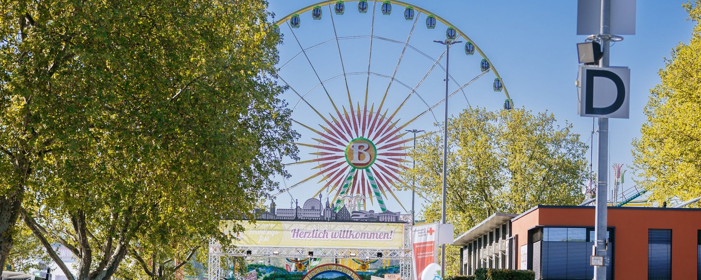 Blick auf Wasen-Eingangstor mit Schriftzug "Herzlich willkommen!". Im Vordergrund Menschen. Im Hintergrund Riesenrad und blauer Himmel.