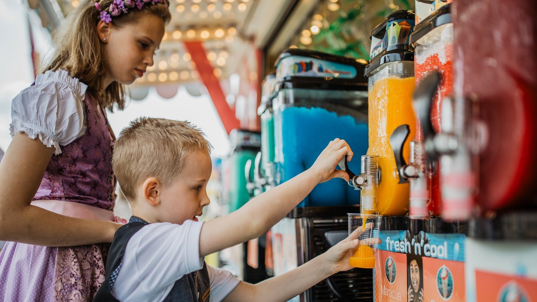 Ein Junge holt sich ein Slush-Eis an einem Zapfhahn. Ein Mädchen in Dirndl steht hinter ihm und schaut dabei zu.