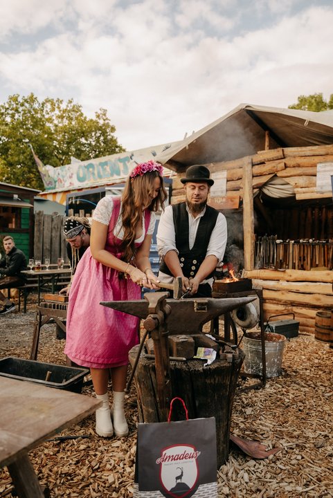 Eine Frau im rosa Dirndl hämmert etwas auf einem Amboss. Neben ihr steht ein Mann im Outfit eines Wandergesellen und hilft ihr.