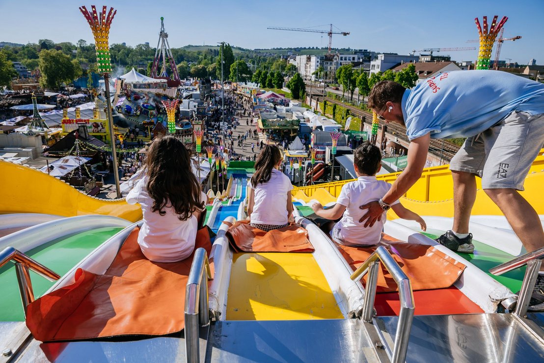 Kinder rutschen auf einer bunten Riesenrutsche nach unten. Ein Mitarbeiter schiebt eines der Kinder an.