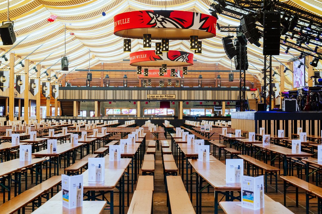 Das leere Göckelesmaier-Festzelt aus der Mitte betrachtet mit Blick auf die Loge an der Pilsbar. Auf allen Tischen stehen Karten. Rechts ist die Bühne mit den Unstrumenten.