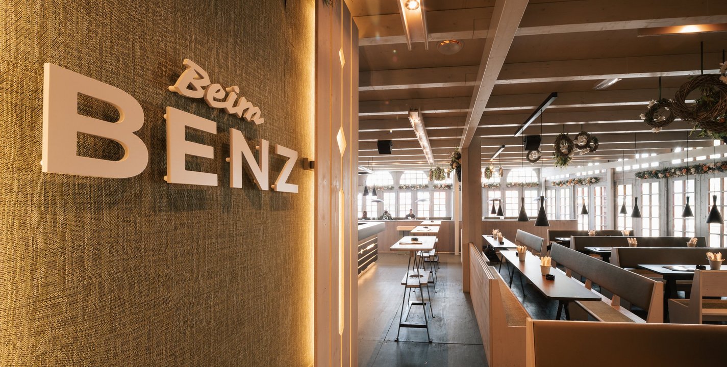 Das Logo vom Festzelt "Beim Benz" an einer Stoffwand montiert. Im Hintergrund sieht man die Bänke und Tische der Loge und ein paar Stehtische.