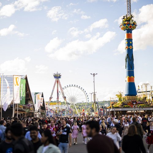 Blick über das Volksfest mit vielen Menschen auf den Wegen. Links stehen die Volksfest-Zelte, rechts thront die Fruchtsäule und in der Ferne sieht man ein Fahrgeschäft und das Riesenrad