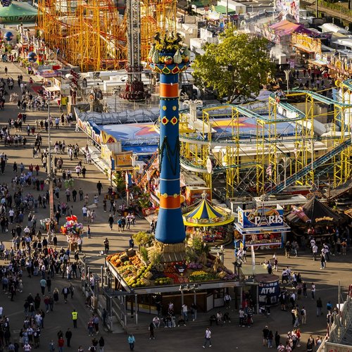 Vogelperspektive auf den Cannstatter Wasen mit der Fruchtsäule in der Mitte. Drumherum sind Fahrgeschäfte und viele Menschen laufen über die Wege und Straßen.