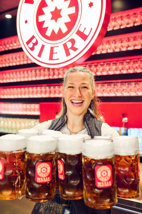 Eine Bedienung hält zehn Maßkrüge und lächelt in die Kamera. Über ihr leuchtet hinter der bar das Logo der Brauerei Wulle.