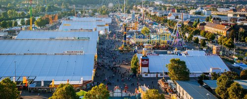Vogelperspektive auf das Volksfest mit den Zelten auf der linken Seite und den Fahrgeschäften auf der rechten Seite. Dazwischen sind unzählige Menschen in den Gassen unterwegs.
