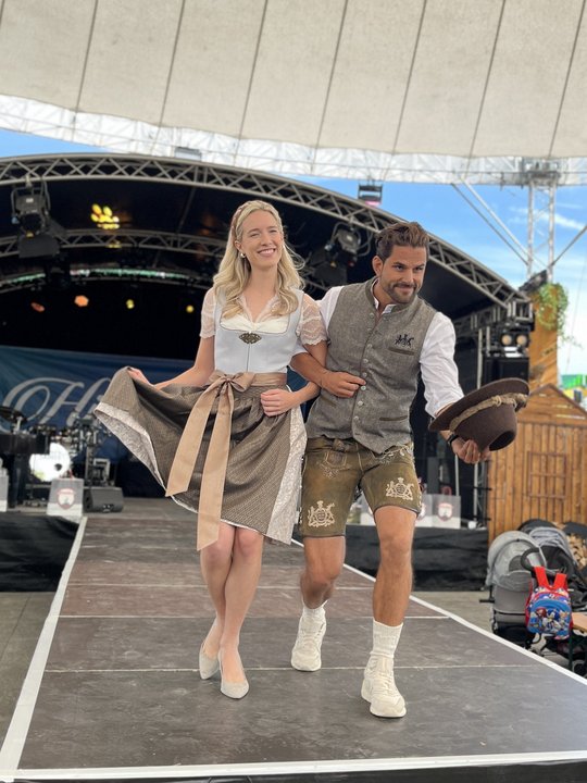 Eine Frau und ein Mann präsentieren auf einer Bühne moderne Trachten und lächeln in die Kamera.