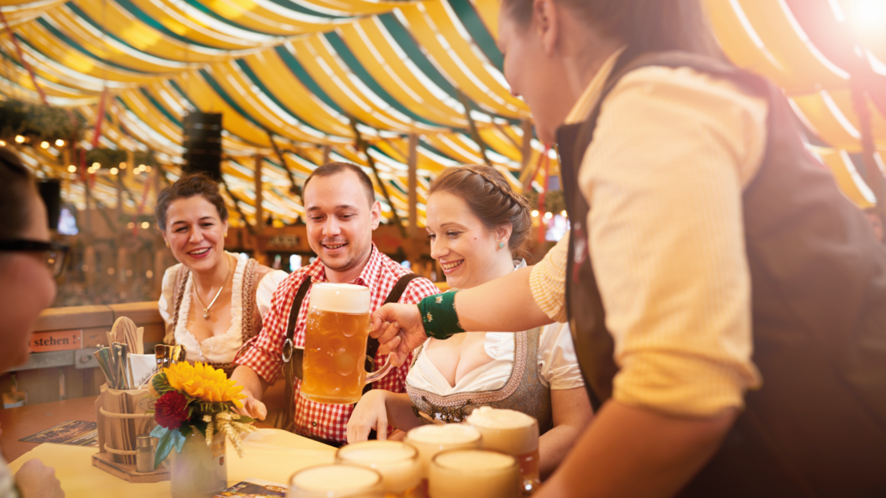 Two women and a man are sitting at a beer table in a tent. A waiter places a beer mug on the table.