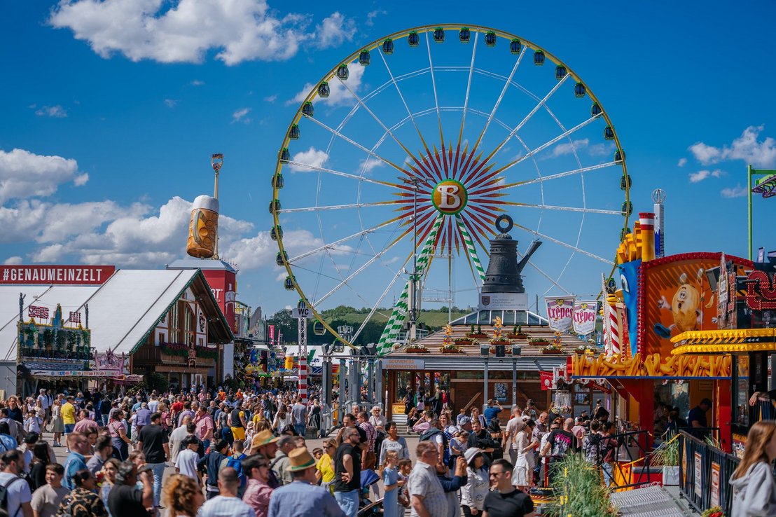 Blick über den Festplatz mit der Cannstatter Kanne auf dem Info-Pavillon. Zahlreiche Menschen laufen über den Platz. Im Hintergrund steht das Riesenrad.