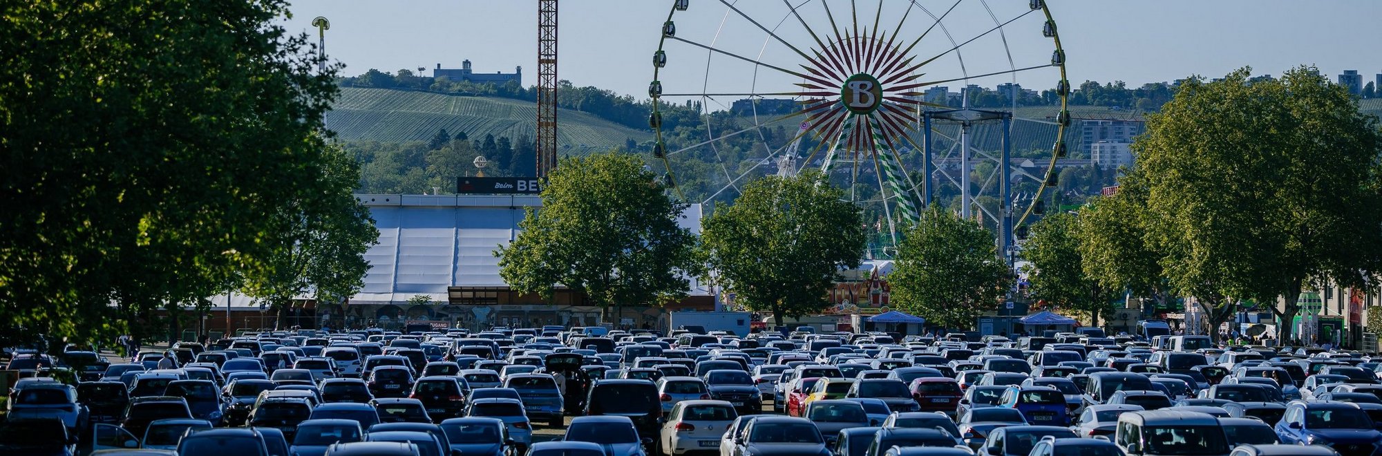 Blick vom Parkplatz P10 auf den Wasen. Im Vordergrund stehen viele Autos, im Hintergrund steht das Riesenrad und das Fahrgeschäft Aeronaut.