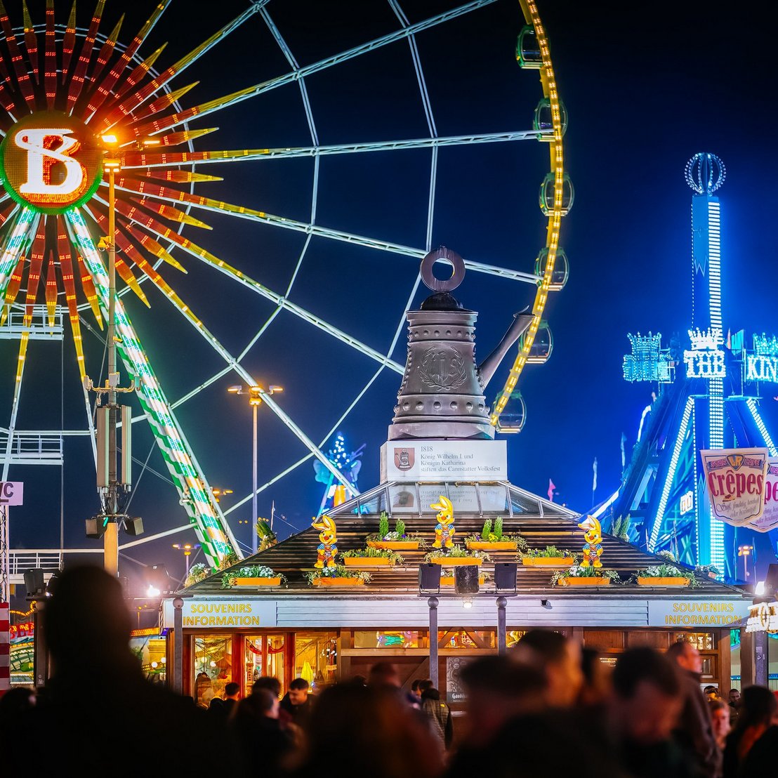 Info-Pavillon mit der Cannstatter Kanne auf dem Dach bei Nacht, dahinter das Riesenrad.