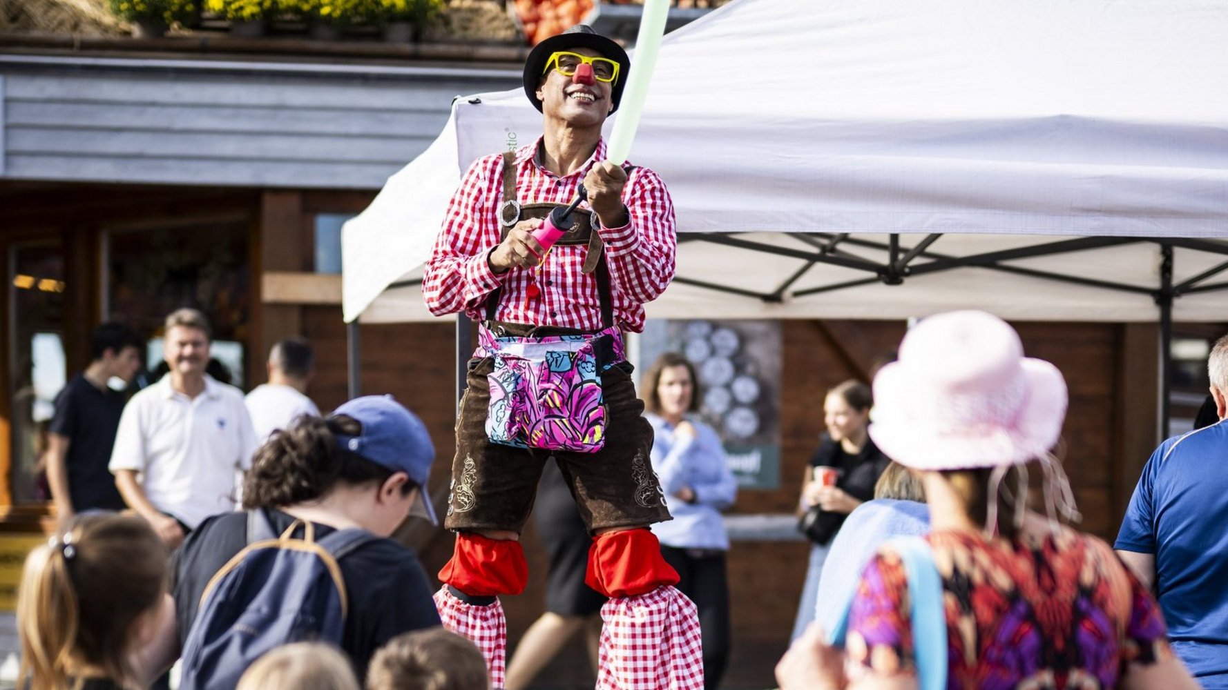 Ein Clown steht auf Stelzen und jongliert. Drumherum stehen Menschen und schauen ihm zu.