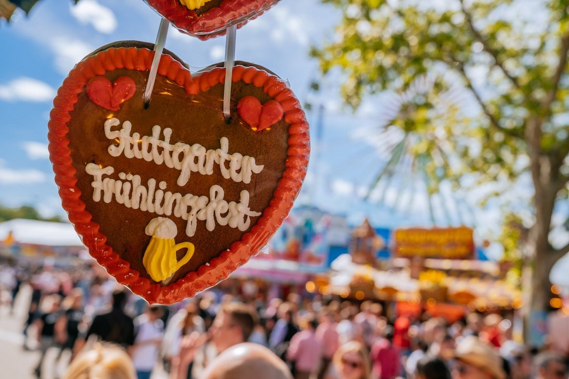 Ein Lebkuchenherz auf dem "Stuttgarter Frühlingsfest" steht. Dahinter ist das bunte Treiben auf dem Fest zu erkennen.