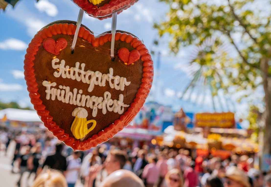 Ein Lebkuchenherz mit der Aufschrift "Stuttgarter Frühlingsfest". Im Hintergrund ist unscharf das Treiben auf dem Fest zu erkennen.