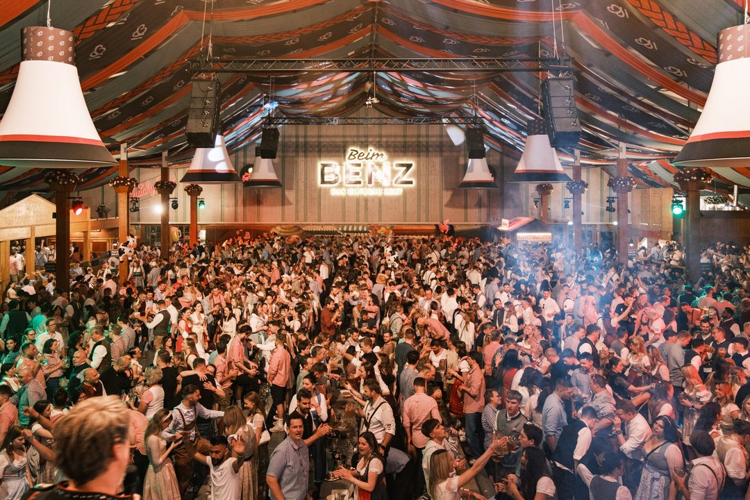 Unzählige Menschen stehen im Festzelt "Beim Benz" auf den Bänken, feiern, singen, trinken und lachen. 
