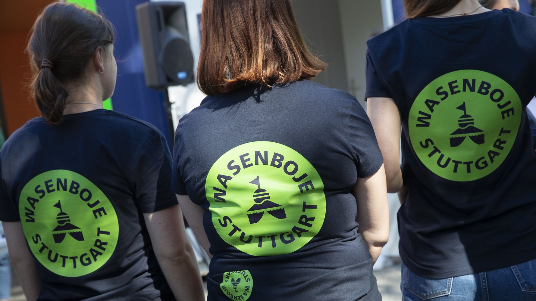 Drei Frauen stehen mit dem Rücken zum Fotografen. Alle haben das gleiche T-Shirt an: ein dunkelblaues Shirt mit dem knallgrünen Logo der Wasenboje Stuttgart