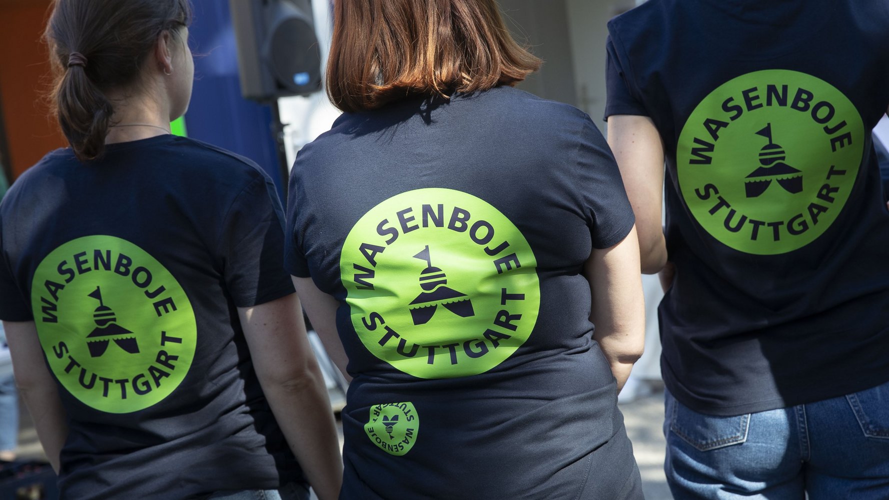 Drei Frauen stehen mit dem Rücken zum Fotografen und haben auf ihren dunklen T-Shirts einen grün leuchtenden Kreis aufgedruckt, in dem "Wasenboje Stuttgart" steht.