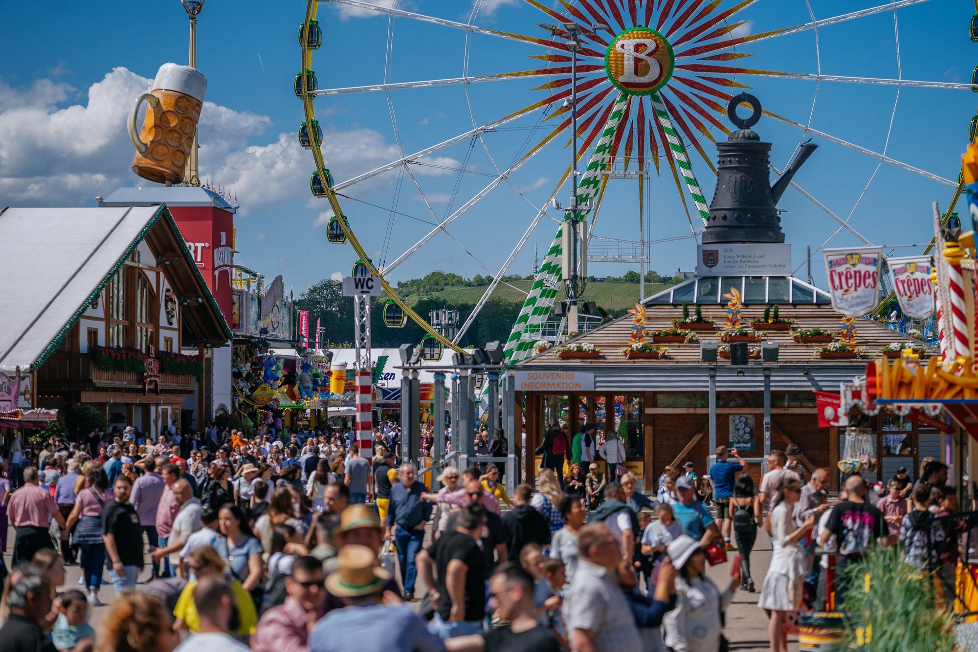 Blick über den Festplatz mit der Cannstatter Kanne auf dem Info-Pavillon. Zahlreiche Menschen laufen über den Platz. Im Hintergrund steht das Riesenrad.